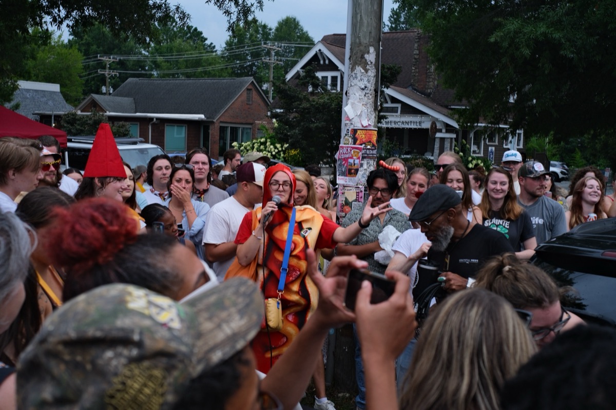 A crowd gathered around a performer in a hot dog costume singing into a microphone on a neighborhood street