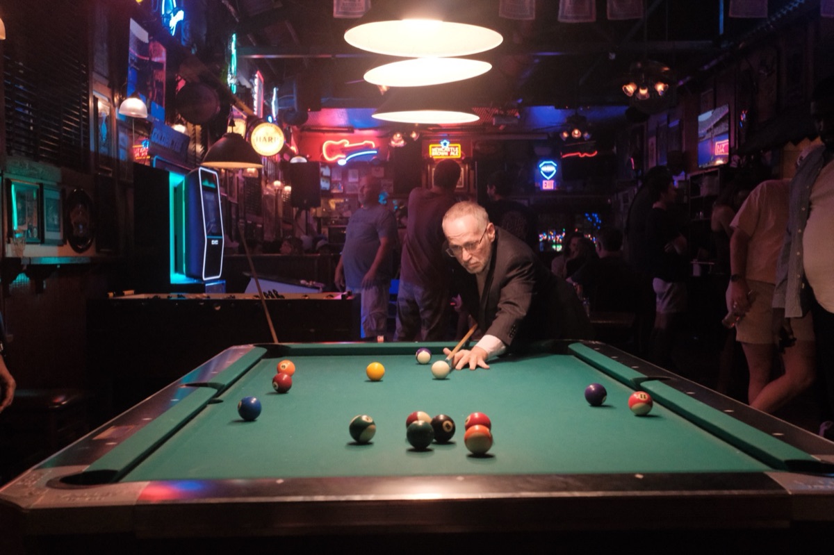 An older man in a dark suit lining up a pool shot in a neon-lit bar