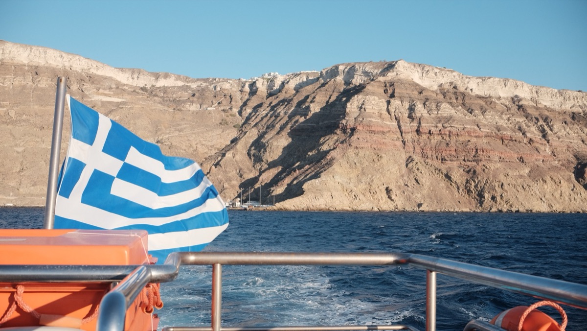 A Greek flag on the stern of a boat, with Santorini's layered caldera cliffs behind
