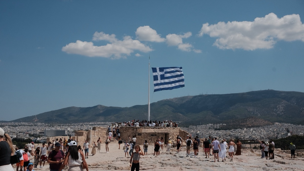 A large Greek flag flying over the Acropolis with the city and mountains beyond